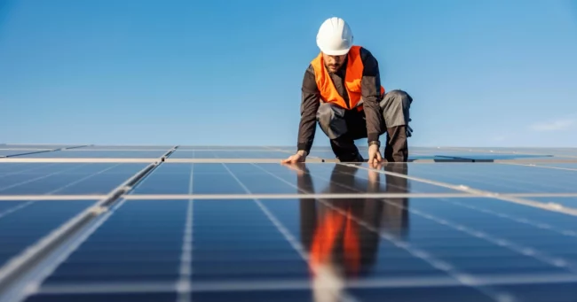 A worker in a hard hat inspects Custom Solar panels in Bluffdale, UT, under a clear sky.